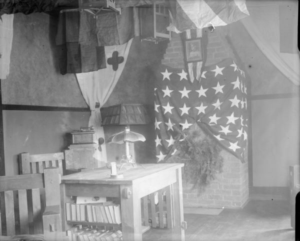 The interior corner of a cabin, showing a number of banners hanging from the ceiling and the fireplace. Other items in the room are a portrait of General John J. Pershing, a wooden table with shelves filled with books, wooden chairs, and lamps.