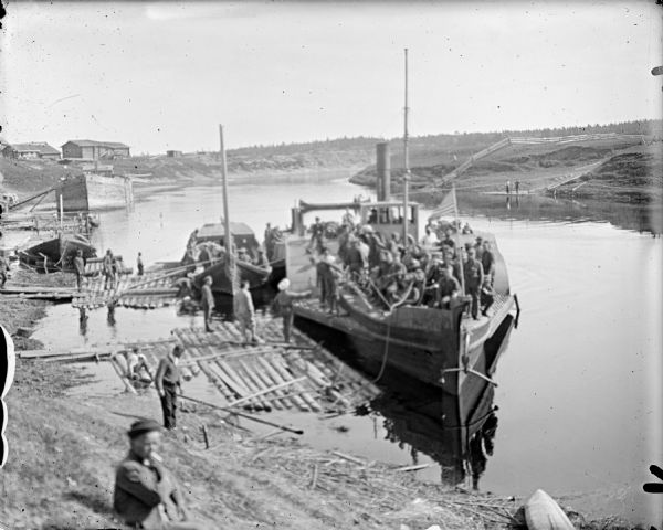 View looking down towards shoreline of troops on board a paddle boat on a canal. The boat flies an American flag. Other people stand on the shoreline and on log rafts floating at the shore.