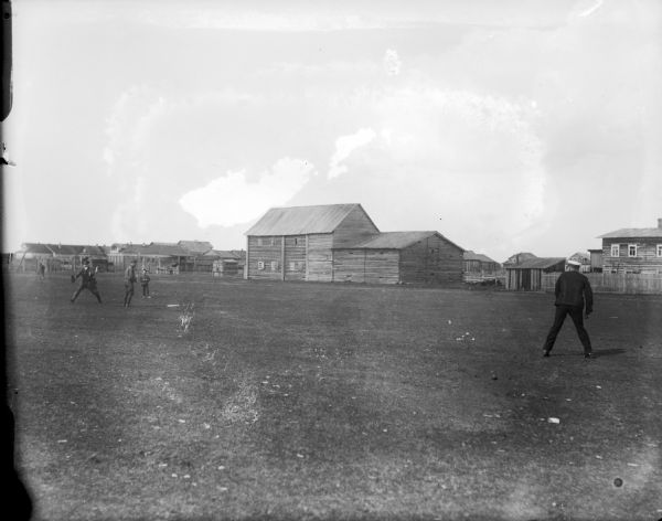 A group of uniformed soldiers playing baseball on the drill field just outside the military barracks. The person pitching the ball is wearing a naval uniform, while the others on the field are wearing army uniforms.