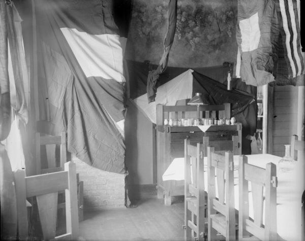 The interior of the officers quarters of the 310th  U.S. Army Engineer Corps while they were deployed to Archangel [Archangelsk], Russia. Flags are hanging from the ceiling, and along the back wall is a small hutch which displays drinking glasses and a table lamp. There are wooden chairs near the window and at a large table which has a glass decanter on a tablecloth.
