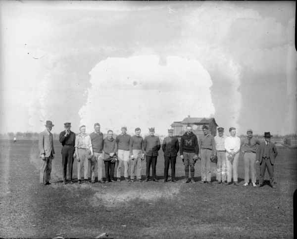 Group portrait of men dressed in civilian clothes, one of which is wearing a University of Michigan hooded sweatshirt. Some of the men have catcher's mitts. Other men are wearing naval uniforms or just a naval cap. In the background is a large wooden building and a fence, possibly enclosing the military drilling field.