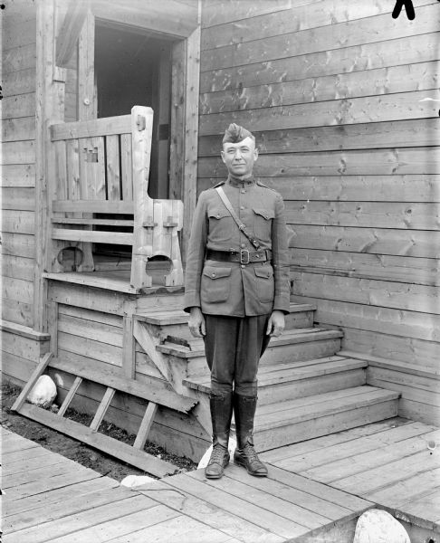 Full-length outdoor portrait of a unidentified uniformed lieutenant colonel or major in the United States Medical Corps standing on a board sidewalk. Just behind him is a wood building with steps leading up to a small front porch with a bench which has the United States Army Engineers emblem carved out of the back.