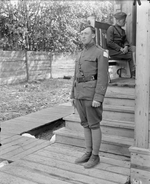 Full-length outdoor portrait of an unidentified uniformed first lieutenant in the United States Medical Corps attached to the 339th Infantry Regiment standing on a board sidewalk just outside a building. He has the shoulder patch insignia of the Army Expeditionary Forces â€” Siberia. In addition, he also has two chevrons on his sleeve, showing one year of active service in the military. The seated soldier sitting on a bench on the porch in the background also appears to be in the Medical Corps.