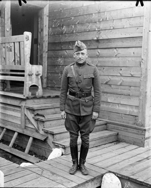 Full-length outdoor portrait of an unidentified uniformed first lieutenant in the United States Medical Corps attached to the 339th Infantry Regiment. He is standing on a board sidewalk, and behind him is a wood building with steps leading up to a small front porch with a bench which has the United States Army Engineers emblem carved out of the back.