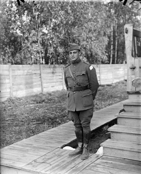 Full-length outdoor portrait of an unidentified uniformed first lieutenant in the United States 310th Engineer Corps. The chevrons on the lower portion of his sleeve, designate that he as seen a year of active overseas military service. He has the shoulder patch insignia of the Army Expeditionary Forces — Siberia on his left. He is standing on a board sidewalk, and just behind him is a wood building with steps leading up to a small front porch with a bench.