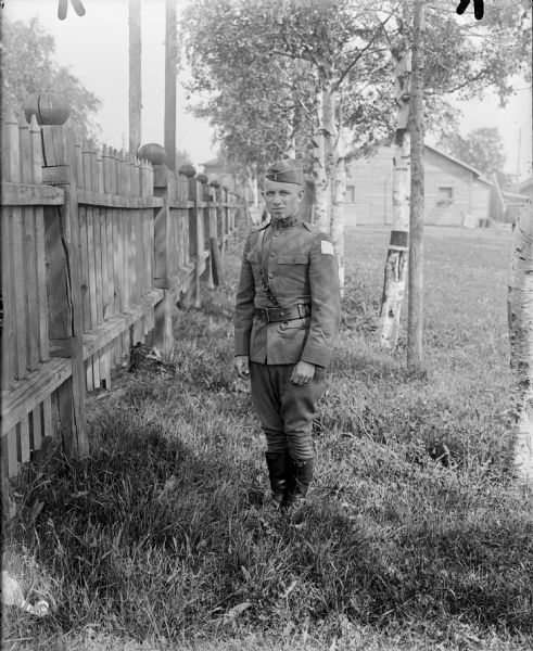 Full-length outdoor portrait of an unidentified uniformed first lieutenant in the United States 310th Engineer Corps standing in the grass next to a fence. The chevrons on the lower portion of his sleeve, designate that he as seen 6 months of active over seas military service. He has the shoulder patch insignia of the Army Expeditionary Forces â€” Siberia on his left.