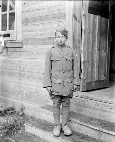 Full-length outdoor portrait of a boy, presumably American, dressed in an American uniform standing on the steps in front of a wooden building. On the child's left shoulder is the patch of the Army Expeditionary Forces - Siberia.