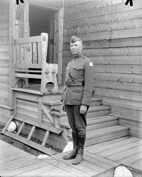 Full-length outdoor portrait of a unidentified uniformed lieutenant colonel or major in the United States Medical Corps standing on a board sidewalk in front of a building which has steps leading up to a small front porch with a bench which has the United States Army Engineers emblem carved out of the back. The chevrons on the lower portion of his sleeve designate that he has seen 6 months of active overseas military service and the shoulder patch appears to be the Army Expeditionary Forces - Siberia.