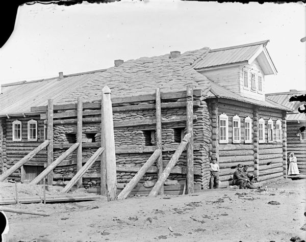 Exterior view of a log building, with sandbags covering a portion of the roof as well as sandbags supported against the wall by wooden posts. Two Russian children and two British soldiers are sitting on the ground at the front of the building. One child is attempting to hide behind the girl in the background.