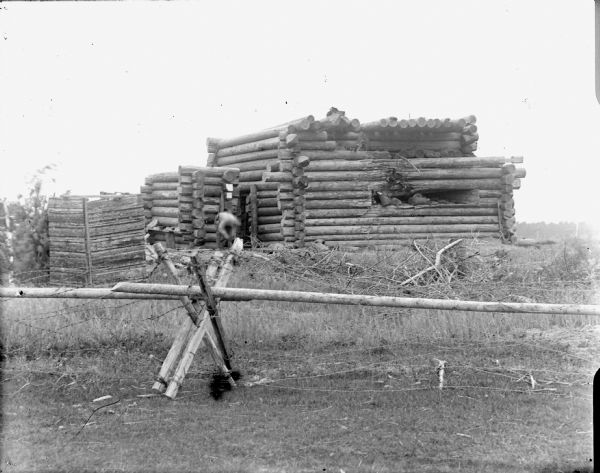 Exterior view of a building constructed of logs that has suffered damage from an attack. The building is behind a wood pole and barbed wire fence. A man near the building (blurred motion) is bending over while working.