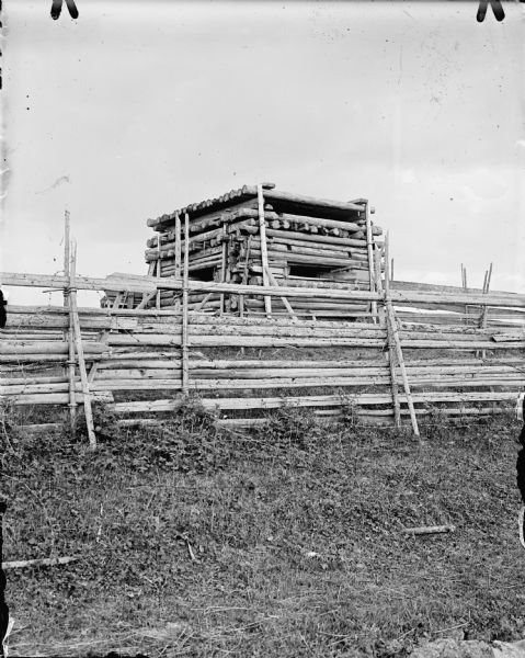 A tall blockhouse constructed of logs behind a wood and barbed wire fence.