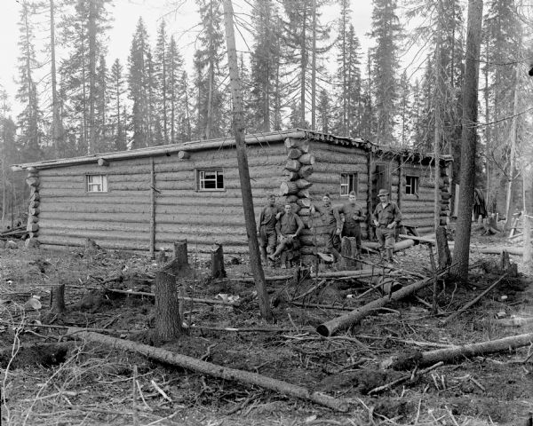 A group of American soldiers posing outside a building constructed of  logs. The area has been cleared of some of the trees, and there are stumps in the foreground.