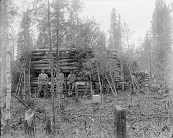 View through stumps and trees of a group of soldiers, possibly French, posing outside a blockhouse constructed of logs.