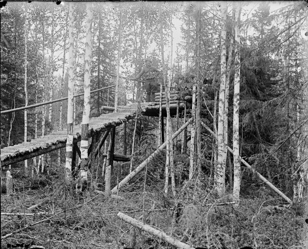 A ramp constructed of logs leads up to a platform in a heavily forested area. An American soldier is standing on the platform.