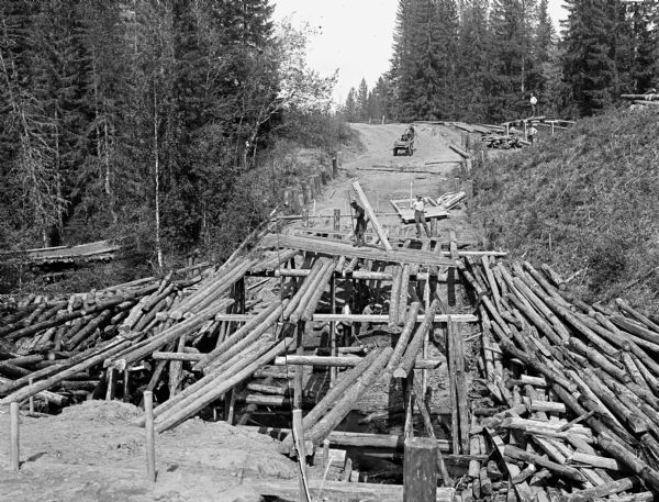 View looking down log bridge which is in the process of being built by a group of soldiers. The bridge crosses over a gully. In the distance is a horse-drawn cart carrying wood supplies. Around the construction site are piles of logs.