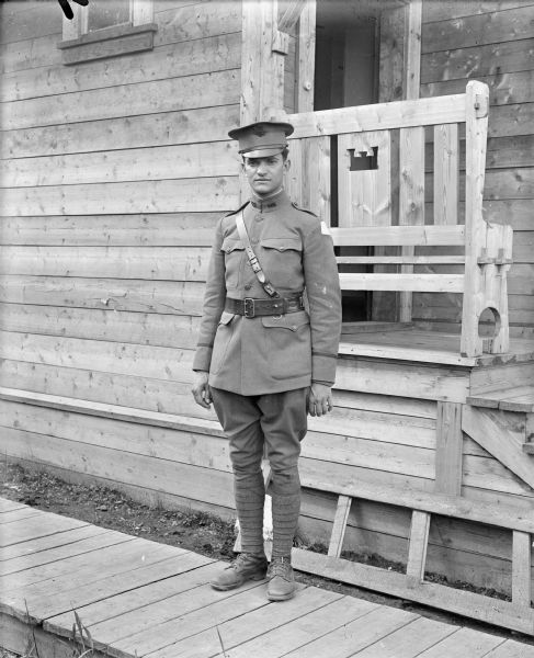 Full-length outdoor portrait of an unidentified uniformed soldier standing on a board sidewalk next to a wood building with a United States Army Engineers emblem carved out of the back of a bench on the small front porch. He has the shoulder patch insignia of the Army Expeditionary Forces - Siberia on his left.