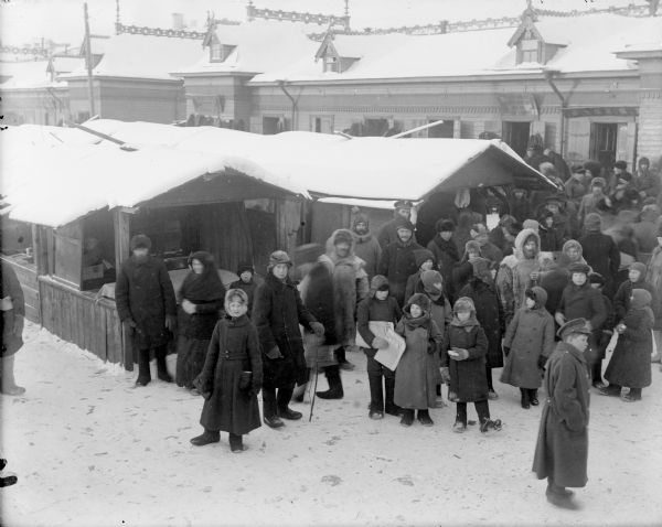 Slightly elevated view of a group of men, women and children standing outdoors in the snow, possibly to attend the market in Archangel [Arkhangelsk], Russia. Amongst the group of people there appear to be a few soldiers, one of which is wearing a cap with the emblem for the British grenadiers.