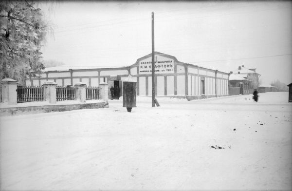 View from street towards the 310th U.S. Army Engineer Corps Supply Depot. There is a small group of soldiers standing in the open doorway of the supply depot under a small sign that says "Engineer Supply Depot." A larger sign above the group on the right is in Russian. There are additional buildings behind the supply depot, and a person walking on the street. On the left is a cast iron fence with columns, and what appears to be a kiosk for signs at the corner.
