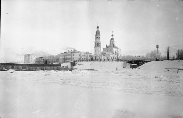 View across snowy ground towards the United States hospital and Army Headquarters for American forces deployed as a part of the Allied Intervention in Northern Russia. On the right is a bridge, and on the left is what appears to be a barge in the ice near pilings. Pedestrians are standing near a railing and one person is standing on what is probably frozen ice below the bridge.