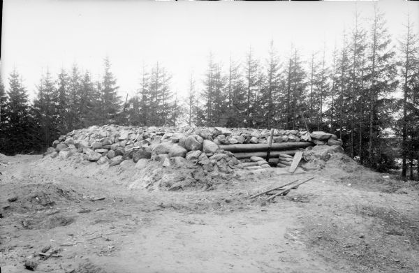 A blockhouse fortification built using a timber frame and large stones, with sandbags stacks near the entrance. There appear to be a wires strung along poles on the far side of the blockhouse.