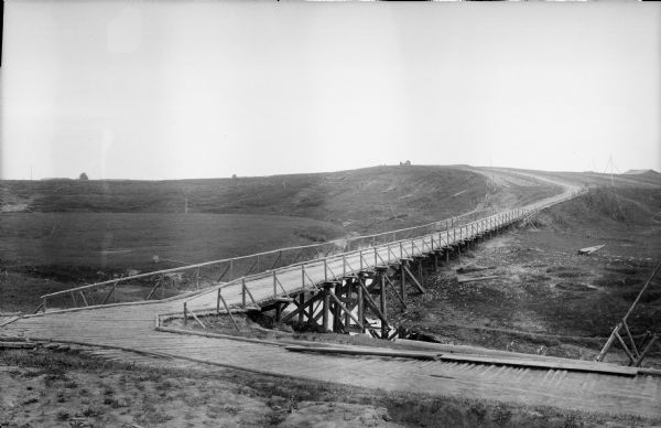 View looking down hill towards a roadbed and bridge constructed out of timbers that cross a small stream or river. In the distance on the far hill is a person leading a horse-drawn cart. On the right, wood poles set up in a tripod have wires suspended over them that lead to a number of buildings over the hill in the background.