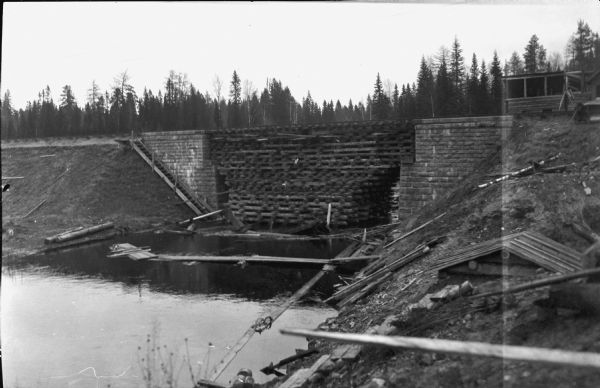 View from shoreline of a reinforced railroad bridge over a river. There are stone foundations on both banks supporting the railroad bed. Wood has been stacked from the river right up to the base of the railroad tracks in order to keep it level. On the right there appears to be a gatehouse. Trees are in the background. A steep set of wooden stairs leads from the railroad bed down to the river. Wood debris litters the river and the bank.