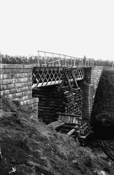 Elevated view from steep shoreline of a railroad bridge repaired with timber stacked all the way up from the riverbed to reinforce the truss of the bridge. There are stone foundations on both banks supporting the railroad bed. On the opposite side of the bridge is a uniformed soldier standing with his rifle on the slope position, controlling the passage of the bridge. The bridges metal railings have been damaged.