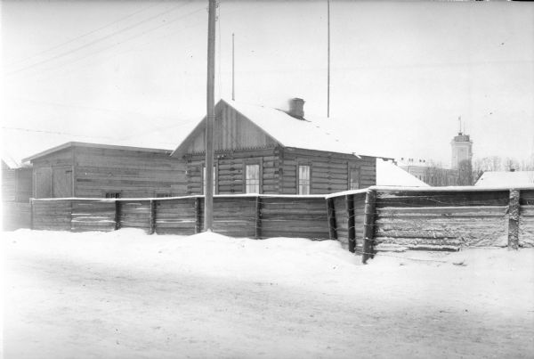View from snowy road of a wooden fence with barbed wire attached to it. Behind it is a building being used as a shed to store lorry (truck) portable radios. Other buildings are on either side. In the background is a building with a tall tower with a clock.