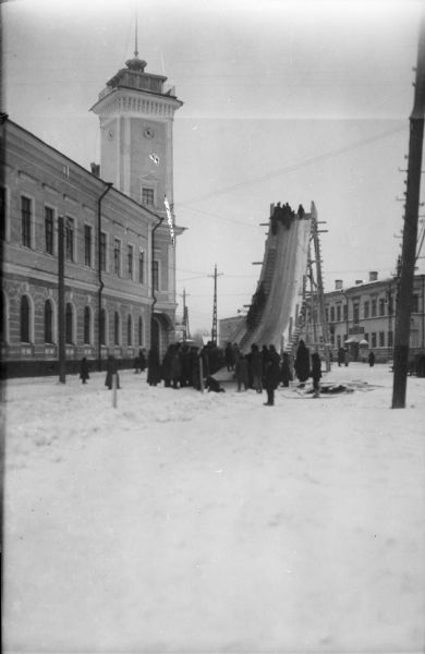 View down snowy road towards a crowd of people gathered at the base of a toboggan slide. A group at the top is preparing to go down the slide. The adjacent building to the left is the Archangel District Administration building.