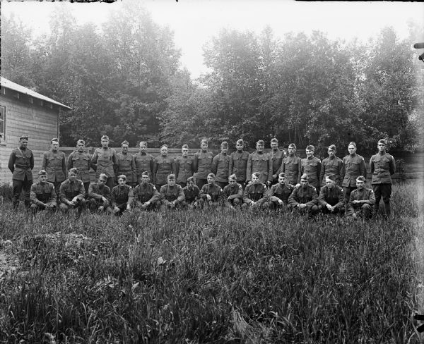 Group portrait of men posing in tall grass in front of a wooden fence. On the left, standing, is a lieutenant, and the rest of the men are sergeants and privates from the 310th United States Army Engineer Corps.