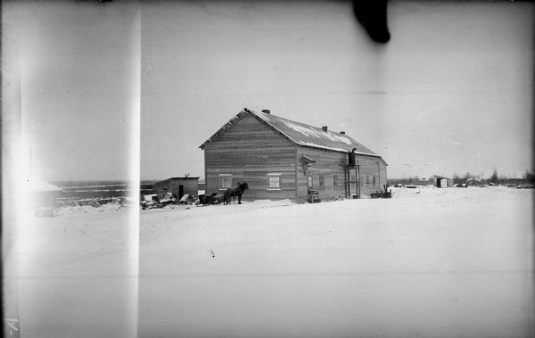 View across snow of a wooden radio station building and a small shed, with other outbuildings in the background on the right. In front of the main building is a horse and sleigh.