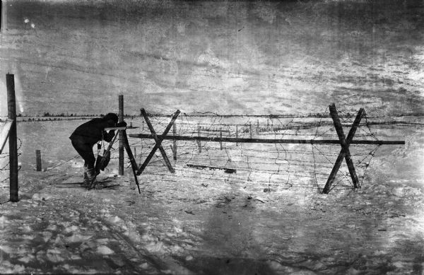 Outdoor view of a U.S. Army Engineer topographer wearing skis while in the process of surveying land. The engineer is standing next to a barbed wire obstacle known as a "knife rest" or a "Spanish rider."