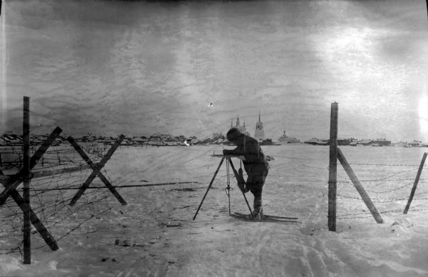 Outdoor view of a U.S. Army Engineer topographer wearing skis while using a tripod stand to write. The engineer is standing next to a barbed wire obstacle known as a "knife rest" or a "Spanish rider." In the distance is a town which appears to be along a shoreline. The most prominent buildings are directly behind the topographer, and appear to be a church and a bell tower.<p