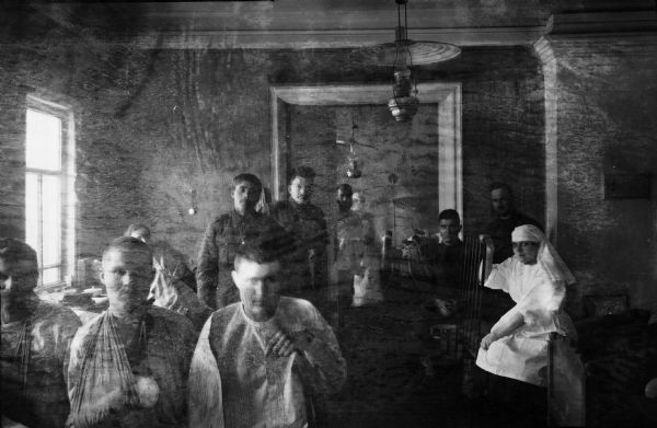 Indoor group portrait of wounded soldiers in the hospital ward, some of which have been wounded. Others soldiers are in uniform and are presumably uninjured. A nurse is sitting on the right on a bed amongst the soldiers. A man is standing in an open doorway in the background. Hanging from the ceiling are oil lamps.