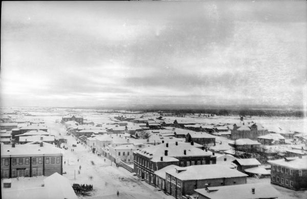Elevated view of Archangel [Archangelsk], Russia. There are pedestrians on the snowy street below, including horses and sleighs. Buildings and dwellings line the streets. In the far background are fields, hills and trees.