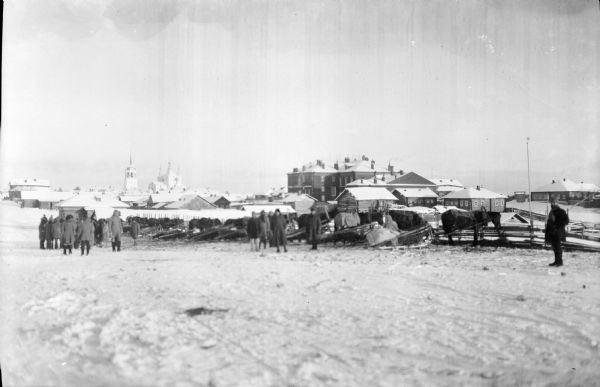 View across snow of a group of men standing around a collection of horses and sleighs that are in front of the quarters of Company G, from the 339th United States Infantry Regiment. In the background is a town, with buildings, dwellings, and what appears to be a church and a bell tower.
