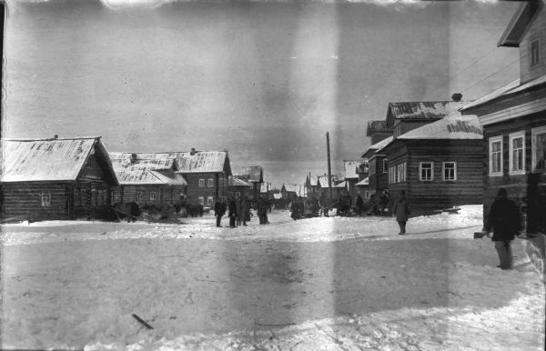 View down snowy street towards a group of people gathered outdoors. Horses with sleighs are parked in front of some of the buildings, which are of log construction.