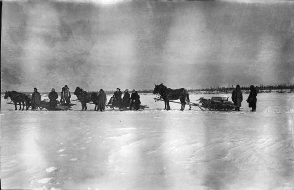 View across snow towards a group of soldiers from the mapping section of the 310th United States Engineer Corps posing with horses and sleighs. In the background are trees along what may be a shoreline.