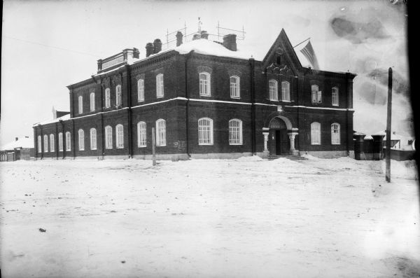 Exterior view across snow of the headquarters building of the United States Army during the Allied Intervention in Northern Russia. An American flag is flying from an open window near the roof of the two-story brick building, which has an arch and two columns framing the entrance.