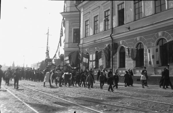 British troops with a regimental band marching into a Russian town. Leading the way ahead of the troops are a group of boys, and closer to the large building walking along the sidewalk are men, and women carrying infants. Tied to the drainage pipes of the building are a series of flags that appear to be maritime signal flags. Streetcar tracks crisscross the street.