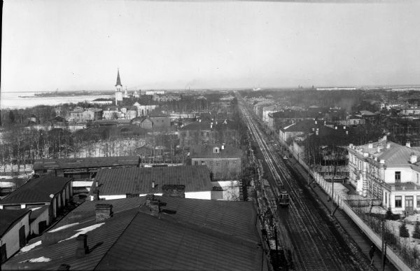 Elevated view of the Troitsky prospect in Archangel [Archangelsk], Russia, looking north. There are dwellings, commercial and industrial buildings, and a church building tower near a shoreline on the left. On the right leading to the horizon is a large street, with a streetcar coming up the tracks.