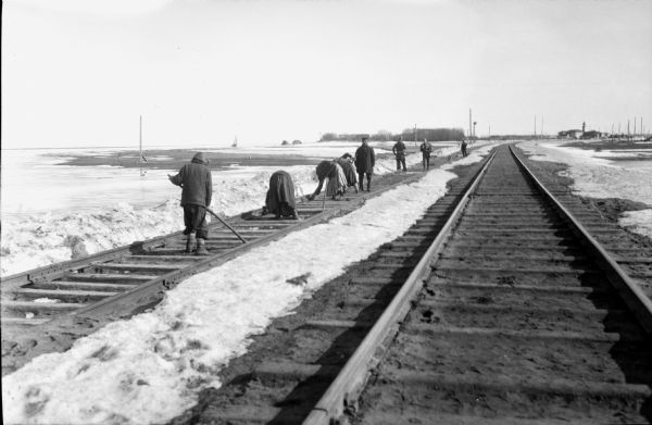 View down railroad tracks toward a group of Russian residents working to clear another set of railroad tracks running alongside. In the far distance are trees and buildings.