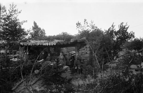 A group of soldiers and officers pose next to an artillery piece within a sheltered area. The shelter is fortified with sandbags and camouflaged with branches from the surrounding area.