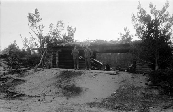 Two Canadian soldiers pose in front of a piece of artillery that is underneath a shelter constructed out of timbers. The shelter is also reinforced with the use of sandbags and camouflaged with branches and fallen trees.