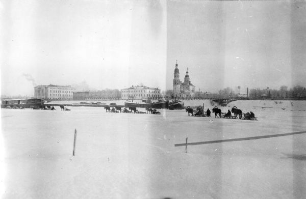 A convoy of horse-drawn sleighs traveling on a frozen body of water. In the background is a church and two other large light-colored buildings, as well as a small truss bridge.
