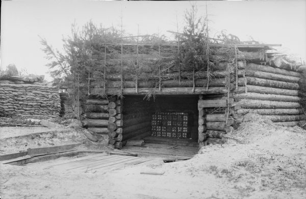 A timber constructed blockhouse housing crates of what is possibly artillery shells. The fortification is camouflaged with branches and fallen trees fastened to the top of the entry into the blockhouse.
