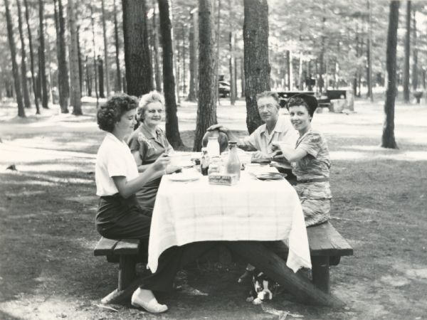 Group of four adults eating at table in picnic area. There is a dog underneath the picnic table.
