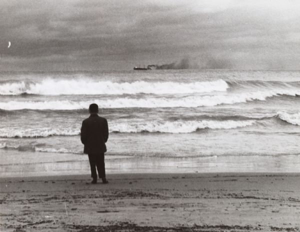 Rear view of a man standing on the shore of what is perhaps Lake Michigan. He is looking out toward moderate breaking surf and a distant freight steamer on the horizon.