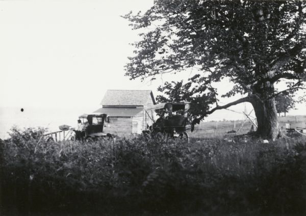 Scene near what may be Coleman, perhaps on the shore of Green Bay on Lake Michigan. View through tall grass towards a man standing next to an automobile looking out towards the lake below. There is a wooden building in the background, and another automobile is parked nearby.
