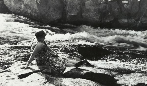 Woman seated on flat rocks, watching rushing water of the Wolf River.